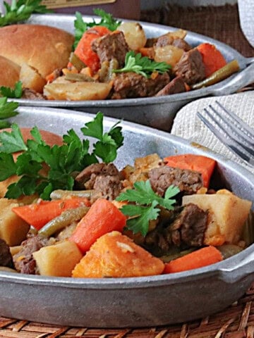 Two comforting looking servings of apple cider beef stew in oval, sliver serving dishes.