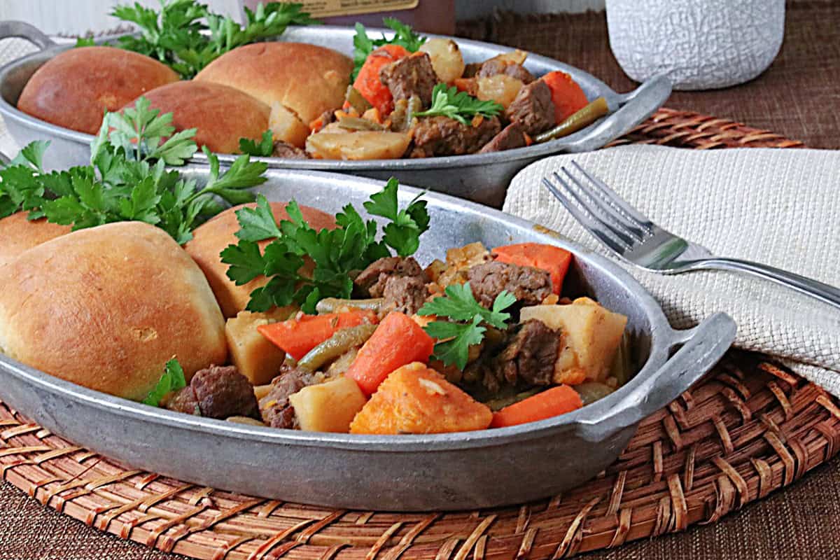 A very homey looking serving of beef stew in a serving dish with rolls, carrots, sweet potatoes, and beef chunks.
