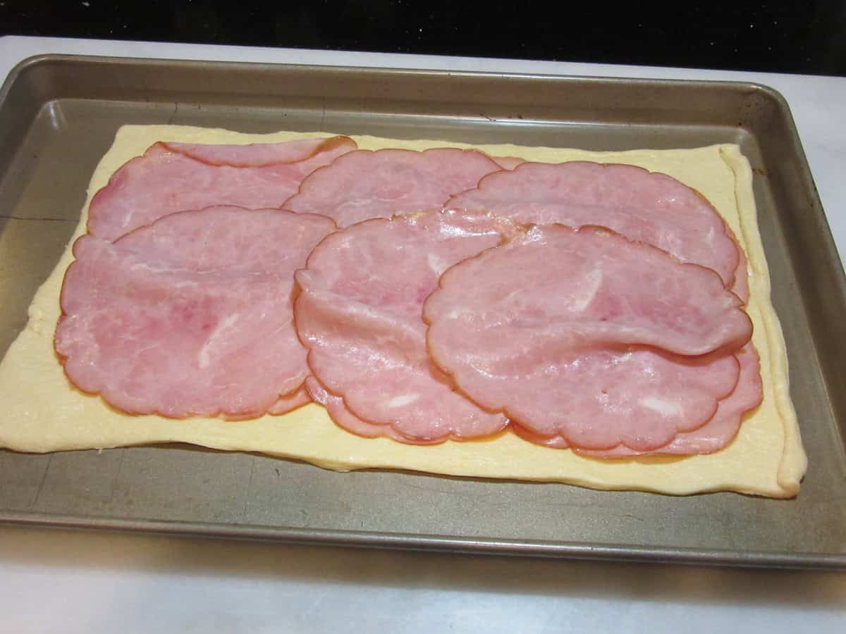 A rectangle of crescent roll dough topped with ham slices on a baking sheet.