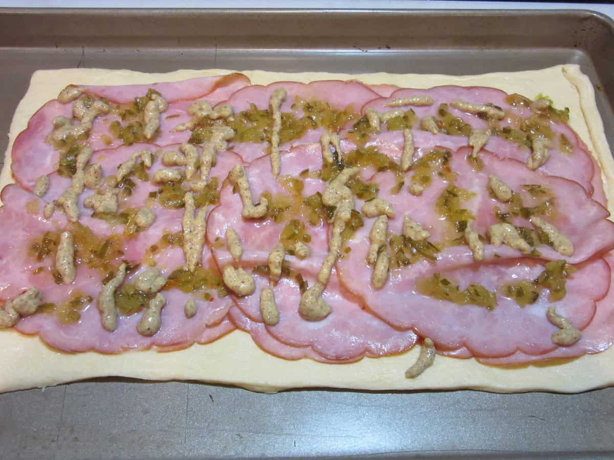 Slices of ham, a drizzle of mustard, and some relish spread out on crescent roll dough.