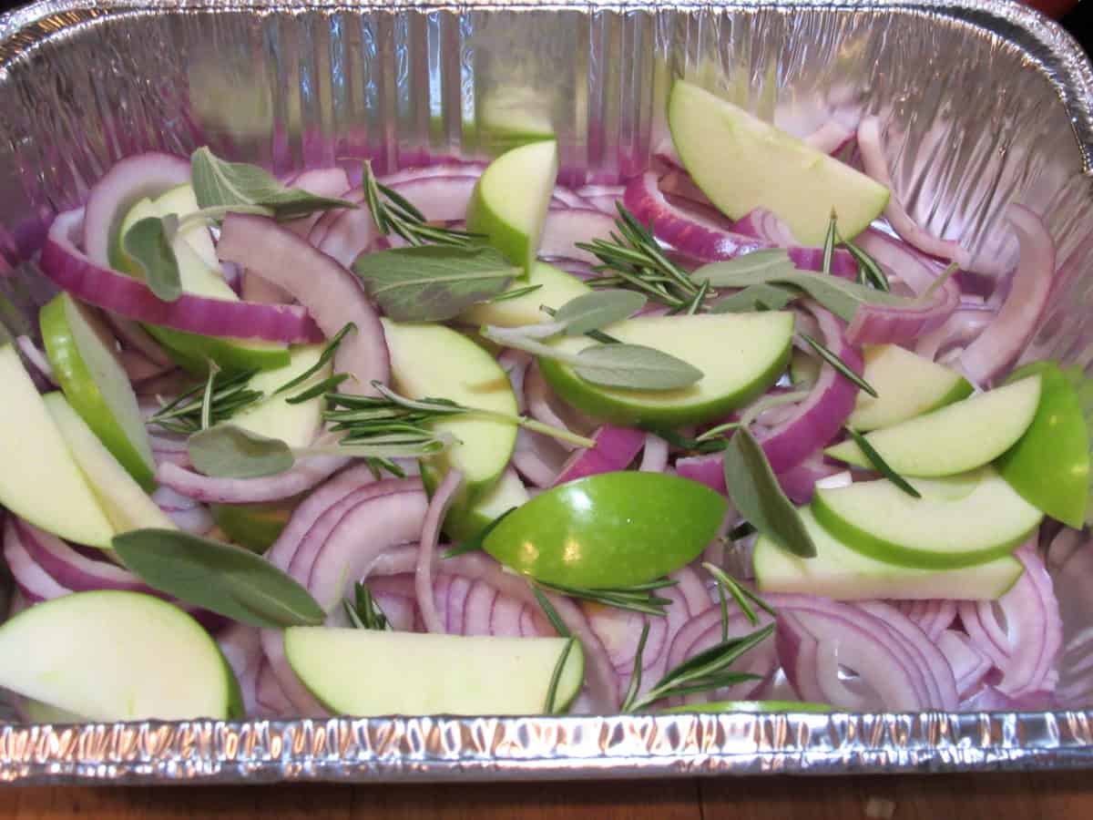 Slices of Granny Smith apples, onions, and herbs in the bottom of an aluminum roasting pan.