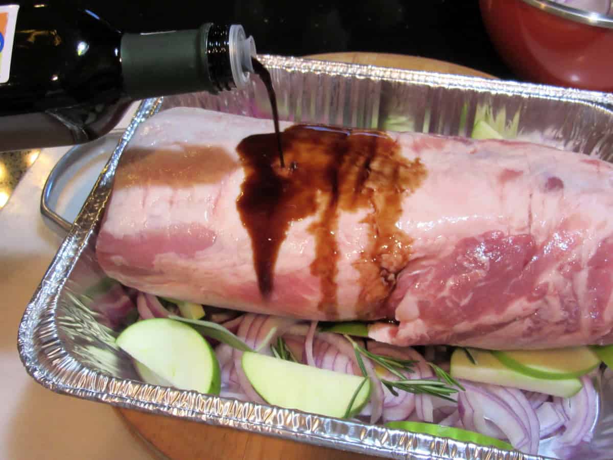 Balsamic vinegar being poured on an uncooked pork roast in a roasting dish.