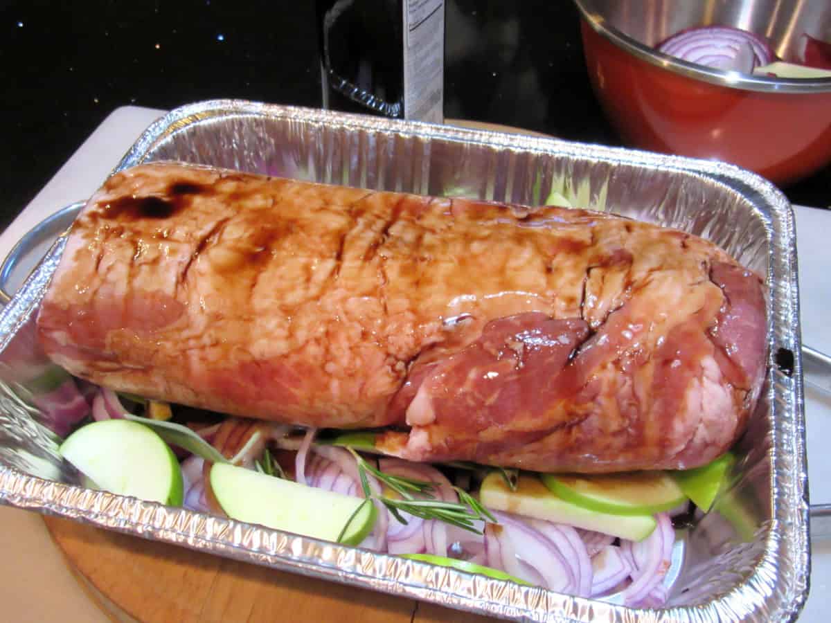 An uncooked pork roast covered with balsamic vinegar in a roasting pan with apples and onions.