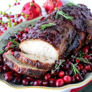 A platter with a cooked cranberry glazed pork roast surrounded by fresh cranberries and herbs.