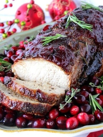 A platter with a cooked cranberry glazed pork roast surrounded by fresh cranberries and herbs.