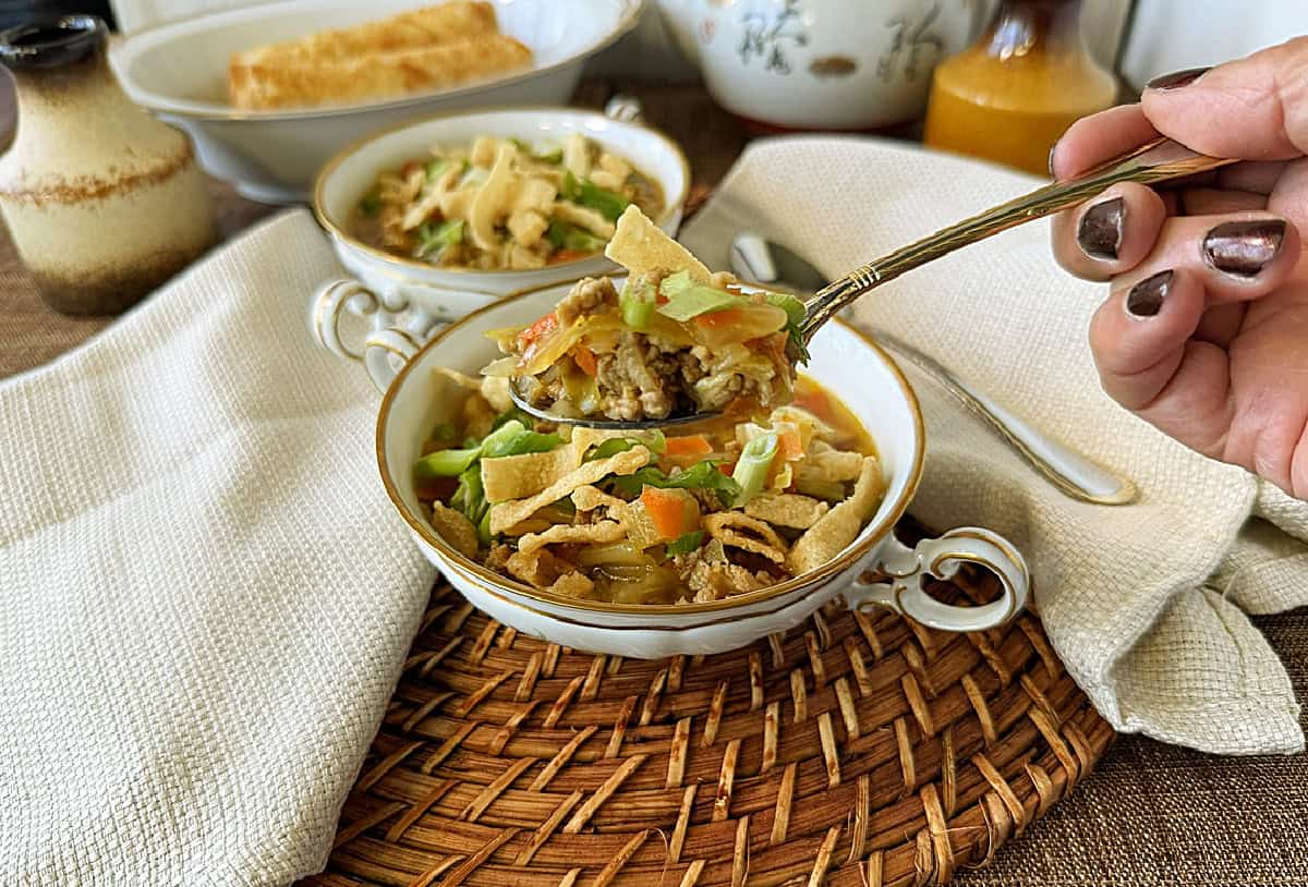 A hand holding a spoon above a bowl of egg roll soup.