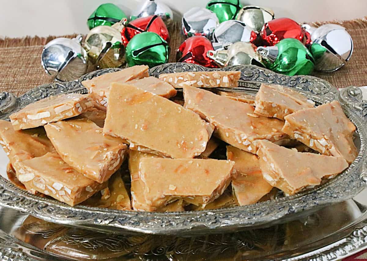 Colorful jingle bells in the background of a bowl of golden Almon Butter Crunch Candy pieces.