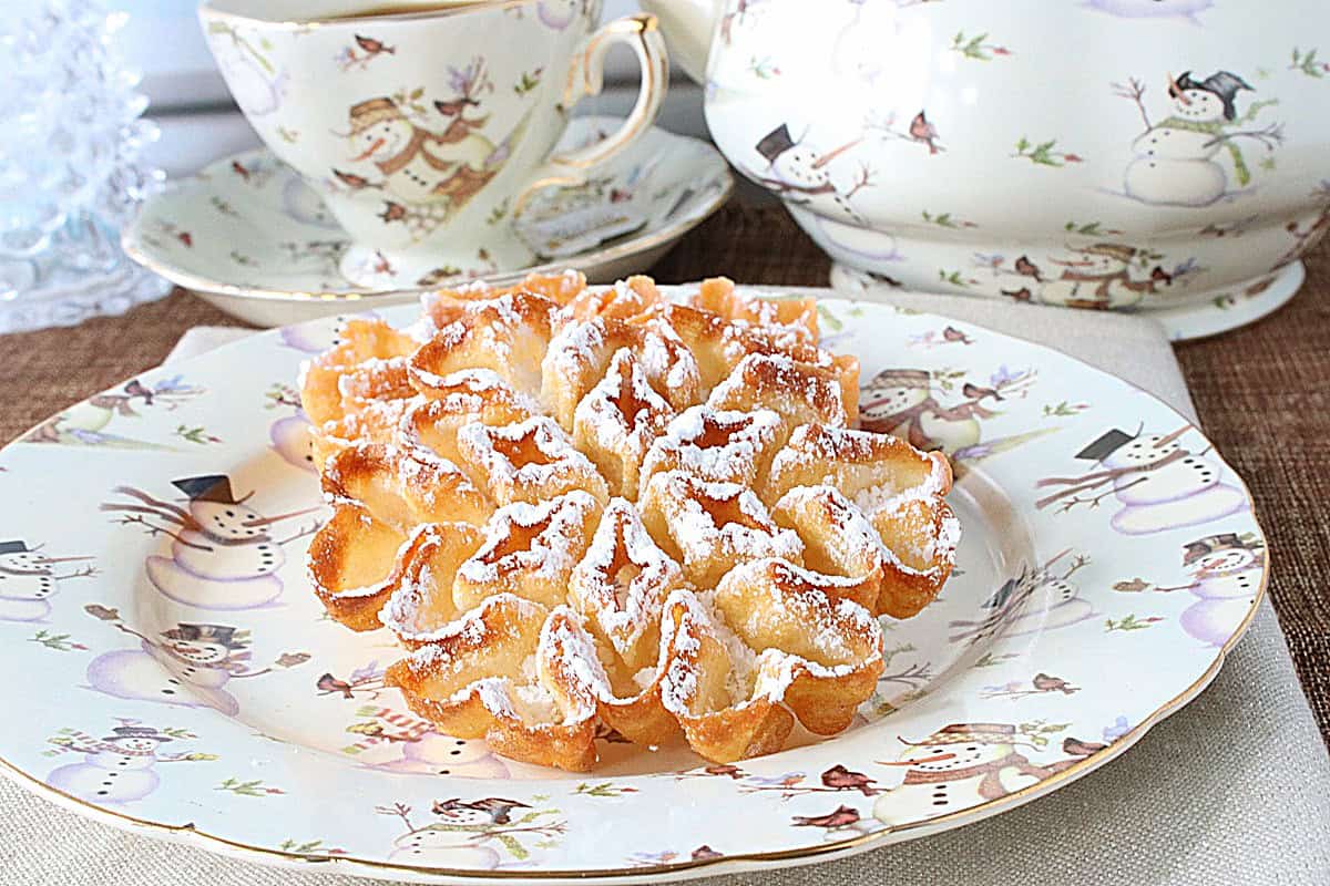 A crispy brown fried rosette snowflake cookie covered with powdered sugar on a cute snowman plate.