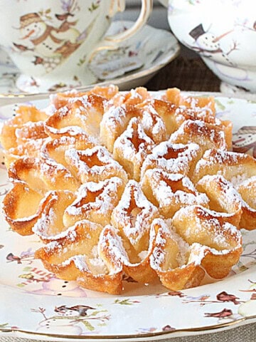 A golden brown fried rosette snowflake dusted with powdered sugar on a plate.
