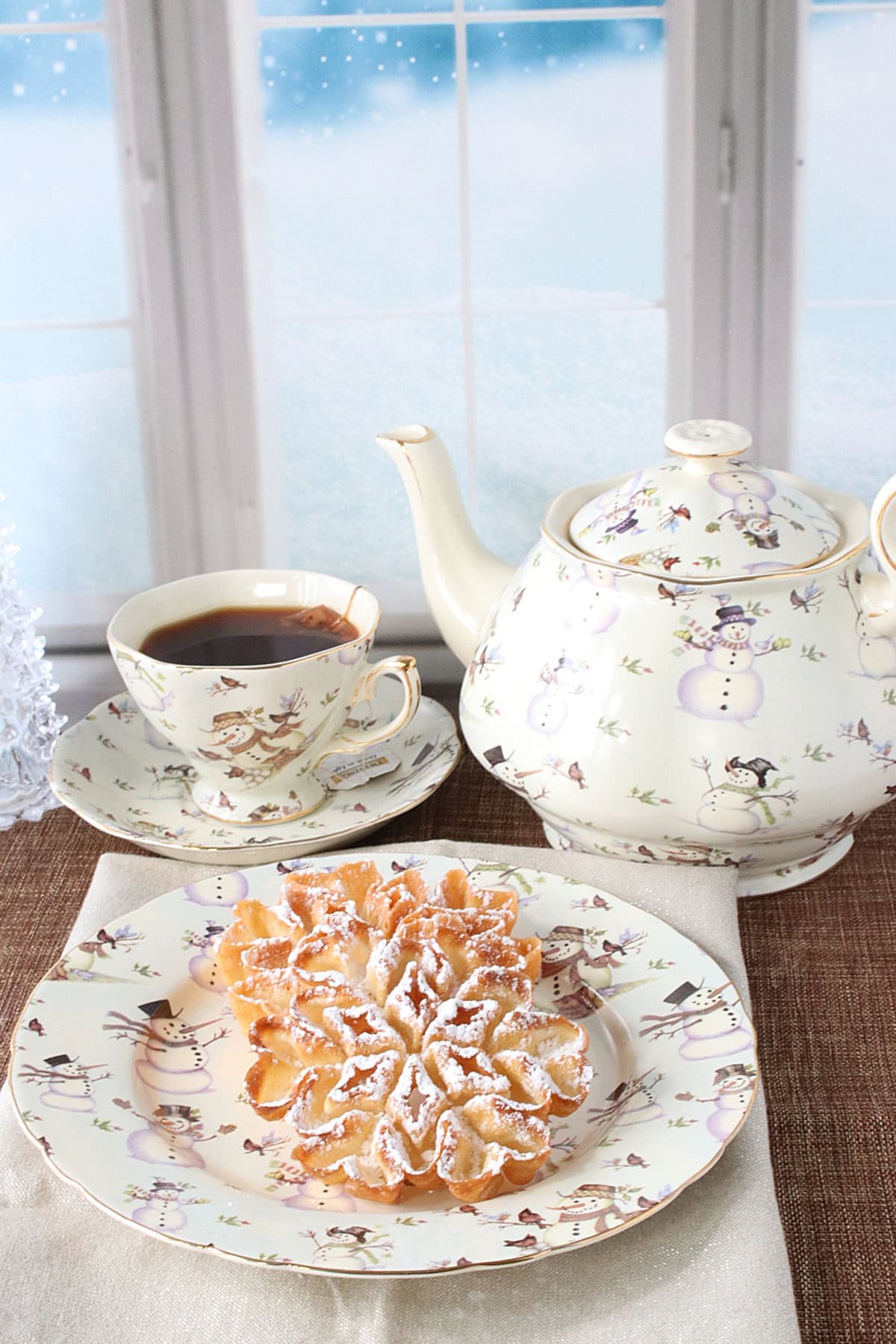 A cute snowman tea pot, cup and plate with a fried rosette snowflake cookie on the plate.