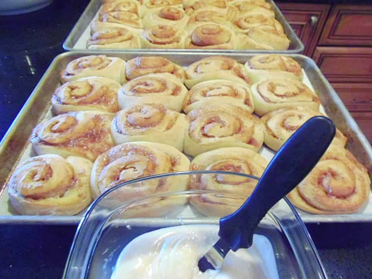 Two baking sheets filled with baked cinnamon rolls and a dish of icing in front.