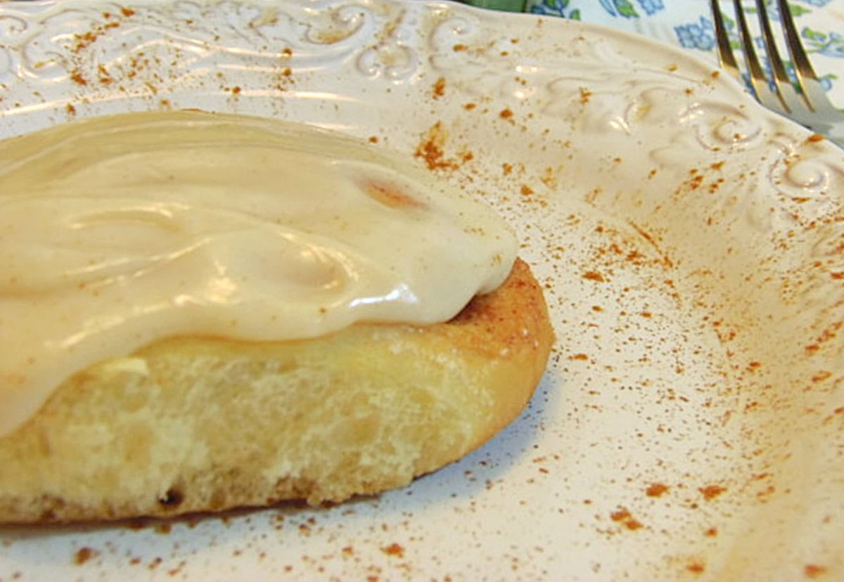 The side view of an iced cinnamon roll on a white plate sprinkled with ground cinnamon.