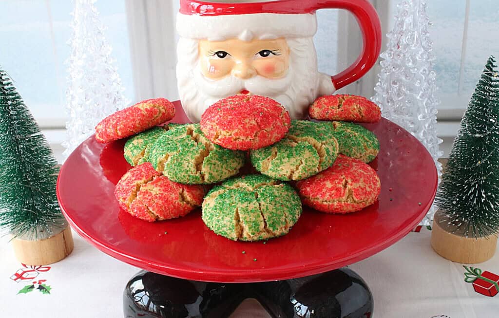 A fun Santa cake plate with a Santa mug on top along with red and green Christmas cookies.