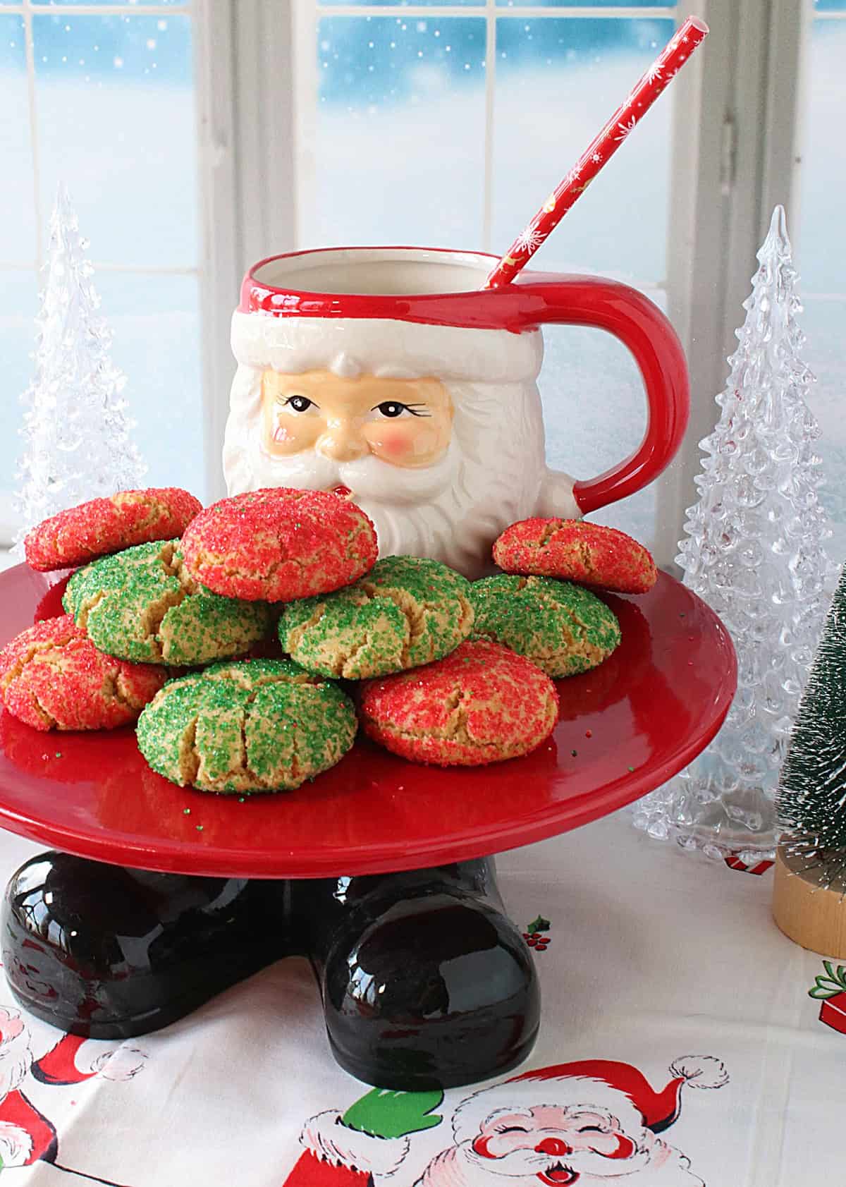 I've been making Santa's Favorite Sugar Cookies every year since I was newly married (36 years ago). And before that, it was my favorite Christmas cookie as a child. It's the cookie that brings back the most wonderful holiday memories for our sons and me, and it's the cookie that we all still consider one of our very favorite Christmas food traditions. - Kudos Kitchen by Renee A Santa cake plate holding Christmas cookies by a window with a winter scene.