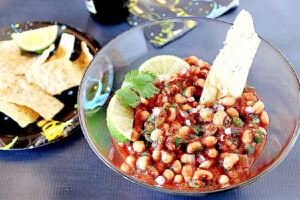 A glass bowl with black eyed pea salsa next to a paper plate with tortilla chips.