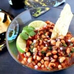 A tortilla chip sticking out of a bowl of black eyed pea salsa with lime wedges on top.