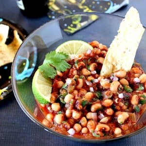 A tortilla chip sticking out of a bowl of black eyed pea salsa with lime wedges on top.
