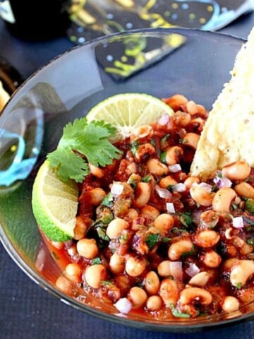 A tortilla chip sticking out of a bowl of black eyed pea salsa with lime wedges on top.