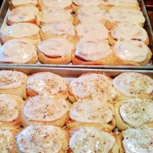 Iced cinnamon rolls on baking sheets with some topped with pecans and some plain.