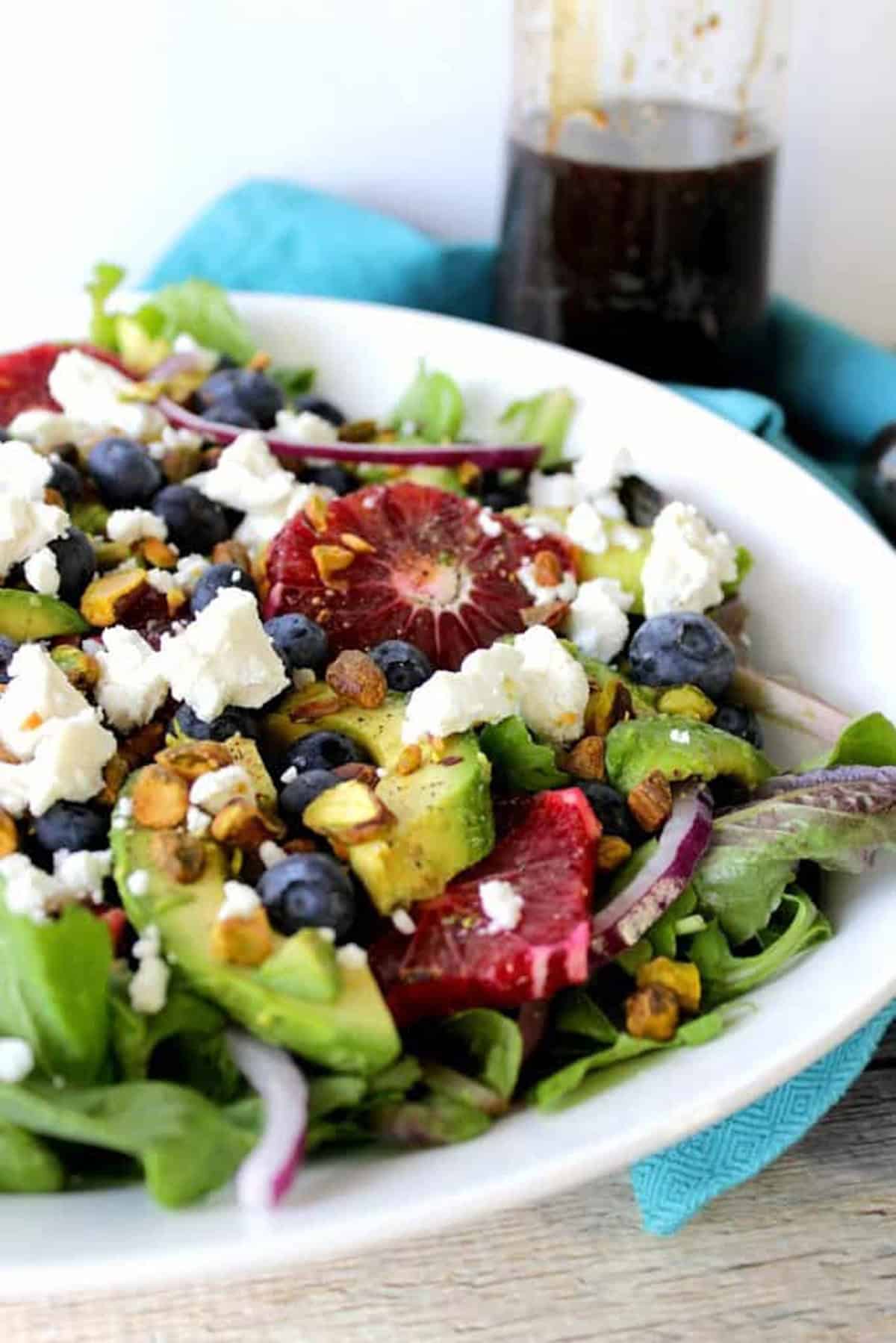 A white salad bowl filled with green lettuces, red onion slices, avocados, blood oranges, and pistachios.