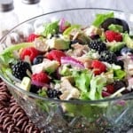 A glass bowl filled with a colorful chicken, berry, and avocado salad along with red onion slices and Parmesan cheese.