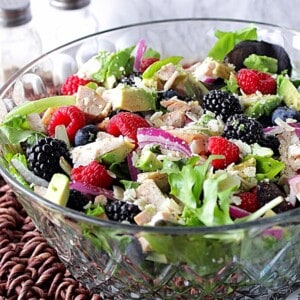 A glass bowl filled with a colorful chicken, berry, and avocado salad along with red onion slices and Parmesan cheese.
