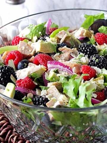 A glass bowl filled with a colorful chicken, berry, and avocado salad along with red onion slices and Parmesan cheese.