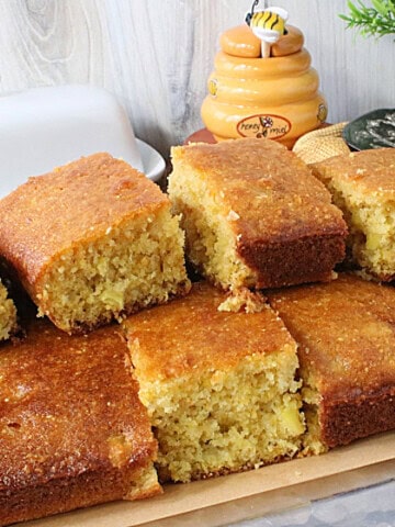A pile of golden brown squares of cornbread with a butter dish and honey jar in the background.