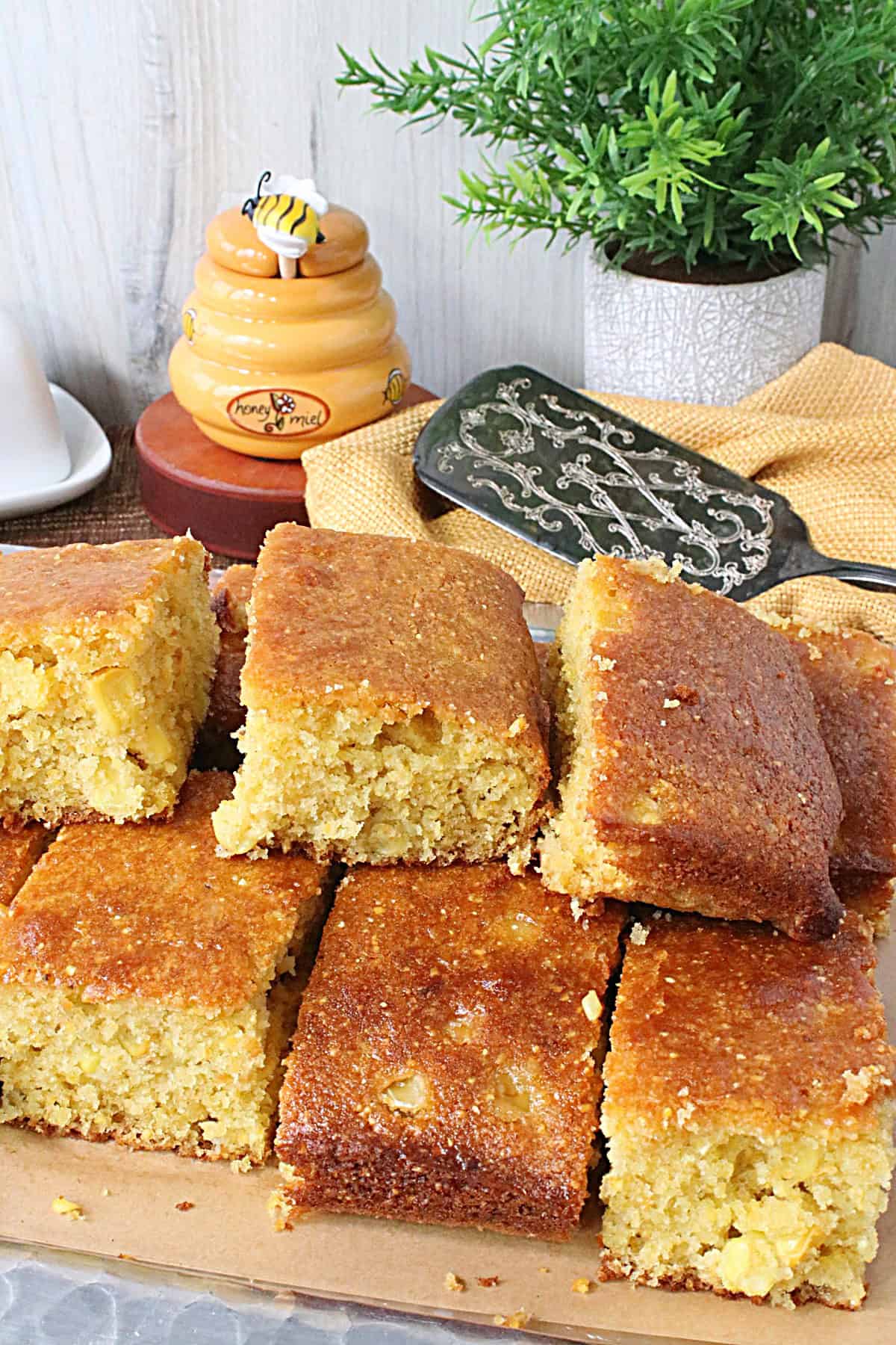 A honey pot and a plant in the background of a platter of perfectly golden cornbread squares.