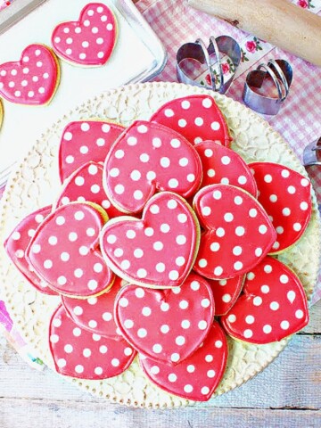A pretty stack of pink polka dot heart decorated sugar cookies on a white cake stand.