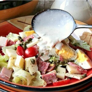 Assorted cheese, meat, herbs, and lettuce in a bowl with blue cheese dressing being poured from a spoon.