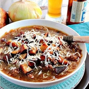 A bowl of German Onion Soup with Bratwurst and Pretzel roll croutons and cheese on top with beer and an onion in the background.