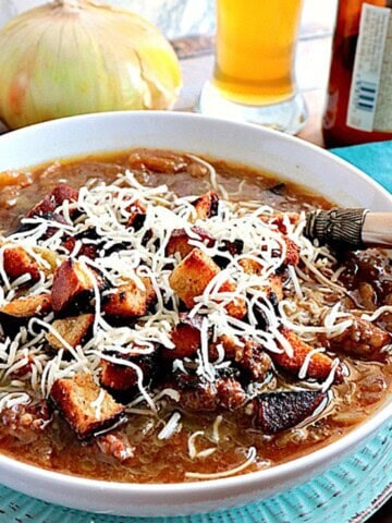 A bowl of German Onion Soup with Bratwurst and Pretzel roll croutons and cheese on top with beer and an onion in the background.
