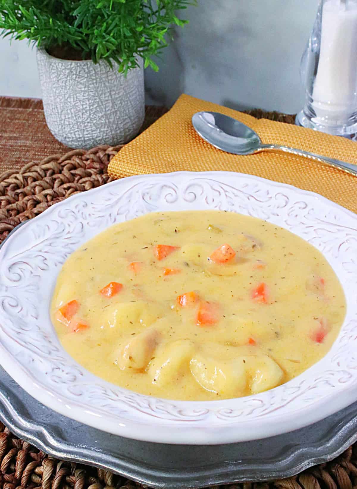 A bowl of cheddar gnocchi soup with a green plant in the background along with a salt grinder and a spoon.