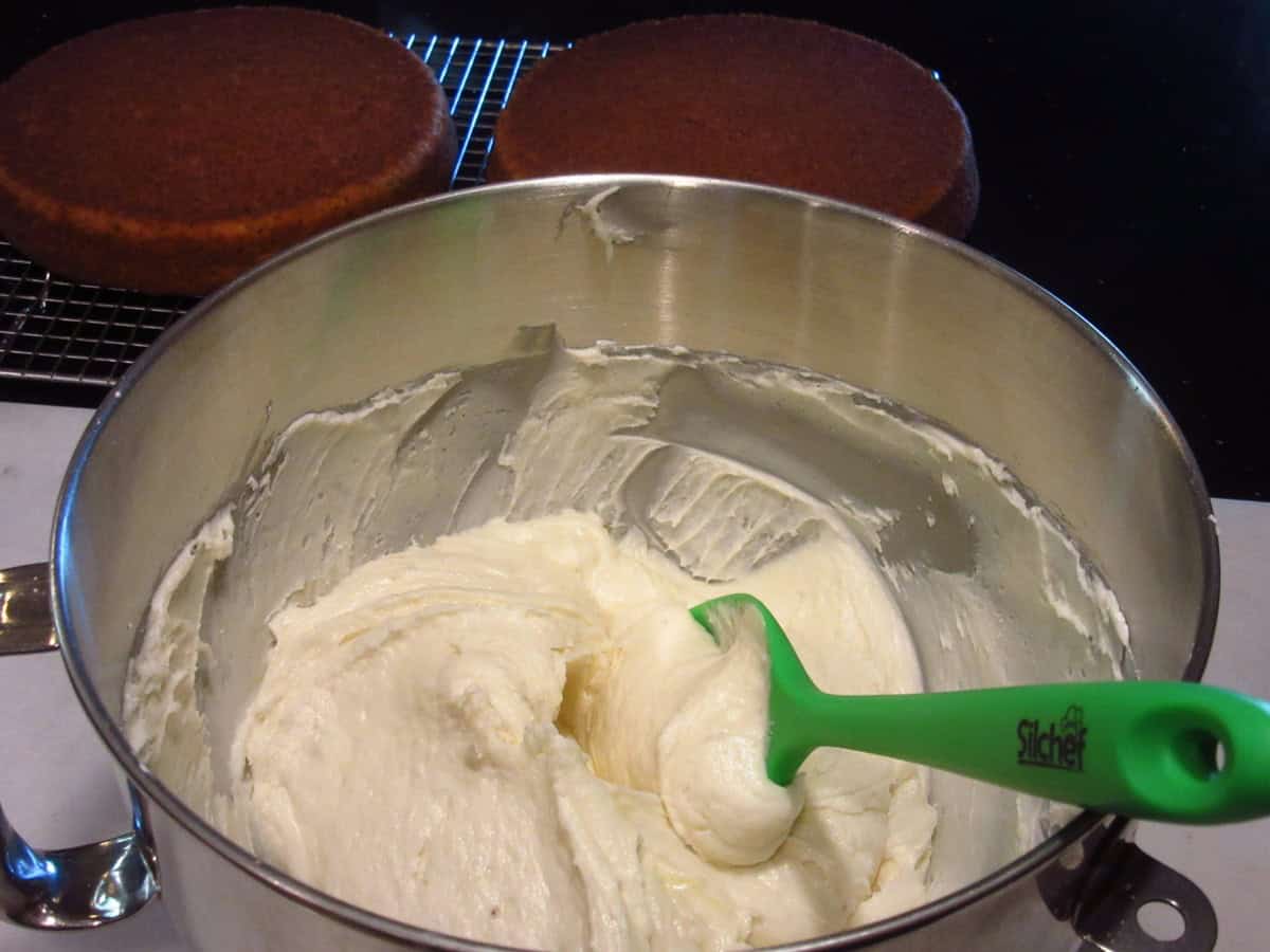 A silver bowl filled with banana buttercream frosting and a green silicone spatula.