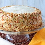 Chopped pecans around a banana buttercream frosted layer cake on a glass cake stand.