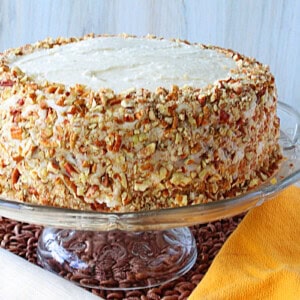 Chopped pecans around a banana buttercream frosted layer cake on a glass cake stand.