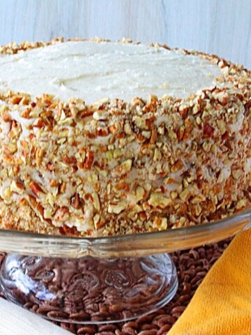 Chopped pecans around a banana buttercream frosted layer cake on a glass cake stand.