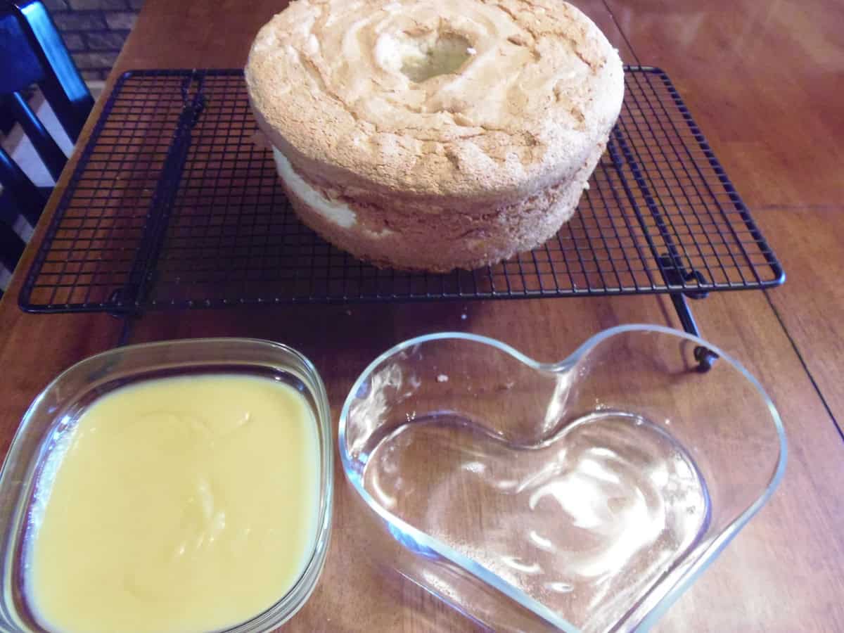 An angel food cake on a wire rack in the background and a bowl of lemon curd and glass heart bowl in the foreground.
