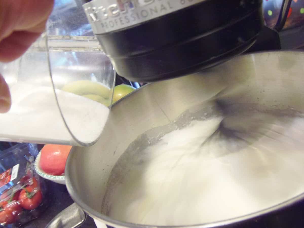 Superfine sugar being added to a stand mixer bowl while whipping egg whites.
