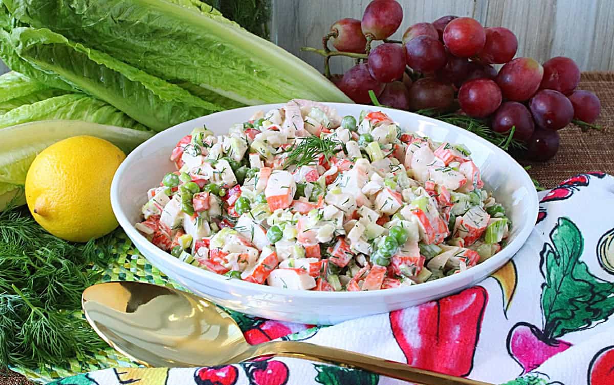 A white bowl filled with crab and pea salad in the center of a colorful napkin, romaine lettuce, and grapes.