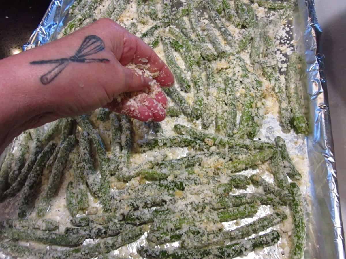 A hand sprinkling potato flakes over green beans on a baking sheet.