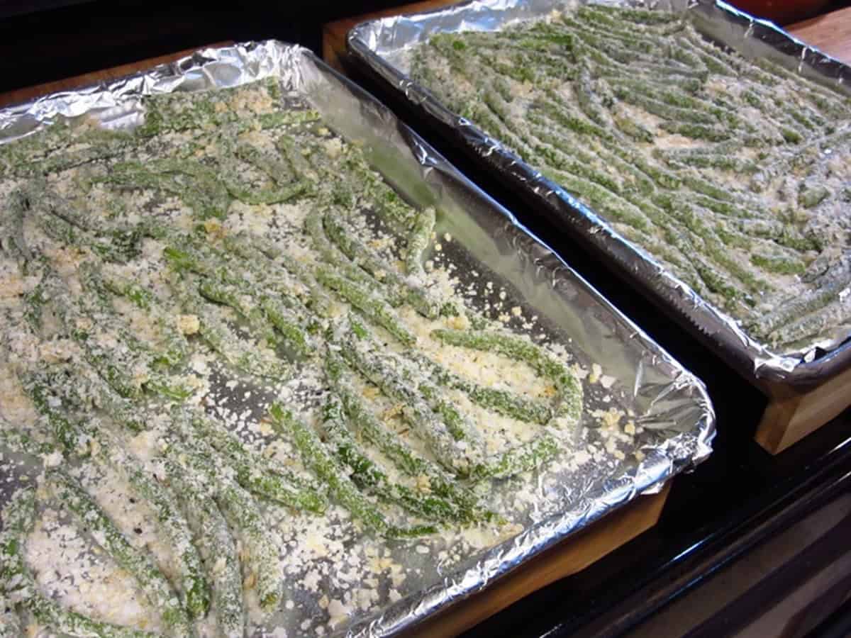 Two baking trays covered with foil and topped with everything green bean fries before baking.