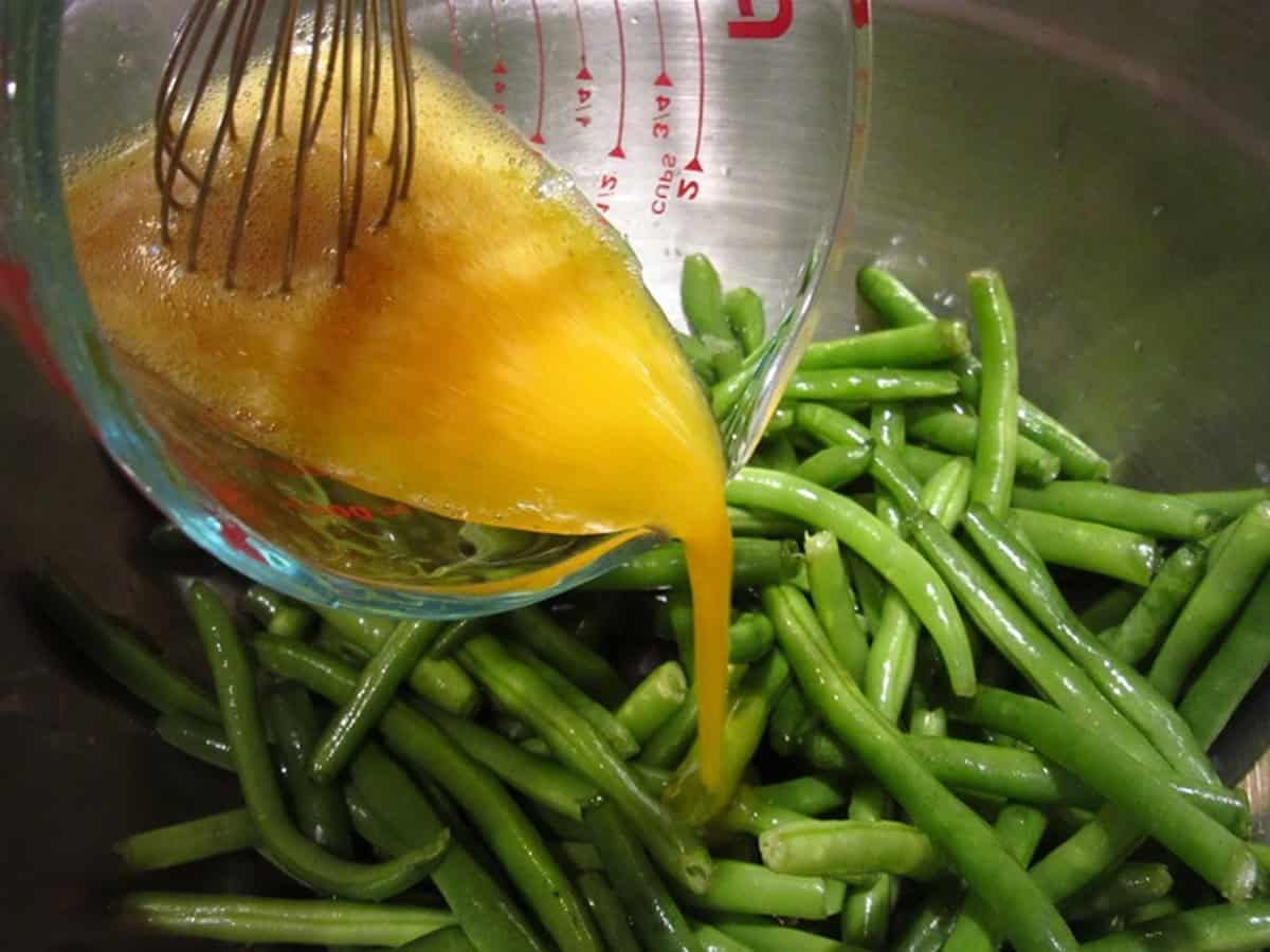 A measuring cup with eggs being poured over green beans in a bowl.