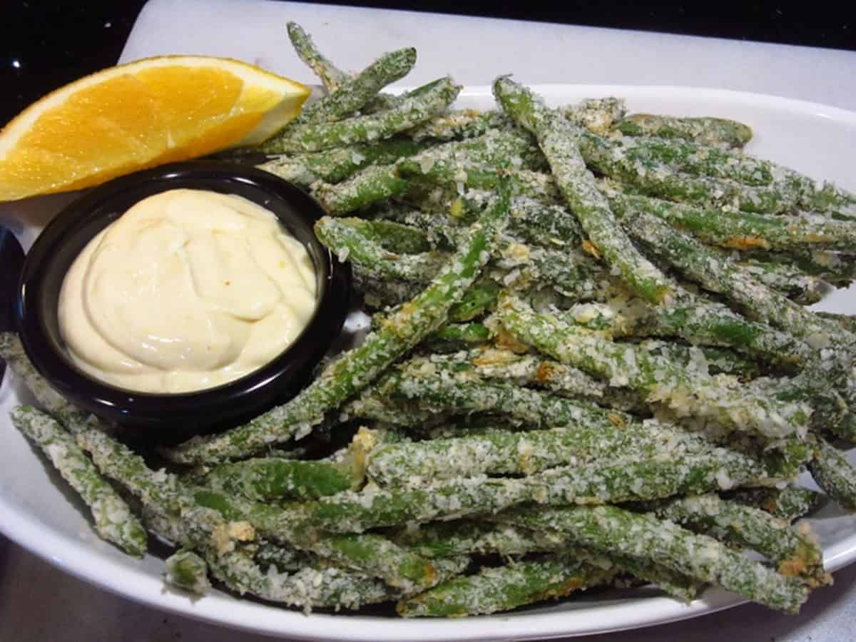 A bowl filled with everything green bean fries along with an orange dipping sauce and an orange slice.