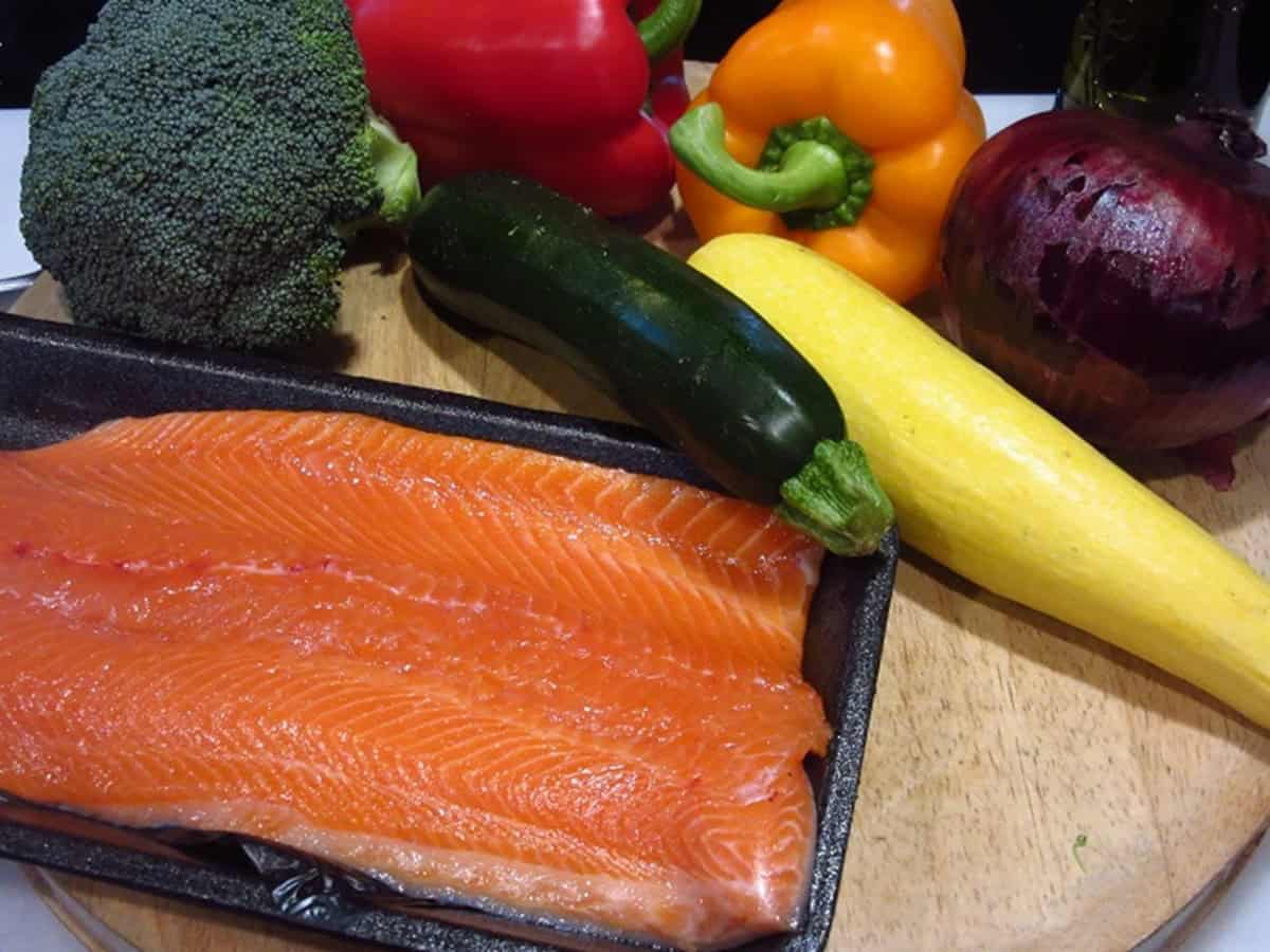 A package of trout surrounded by colorful vegetables on a wooden cutting board.