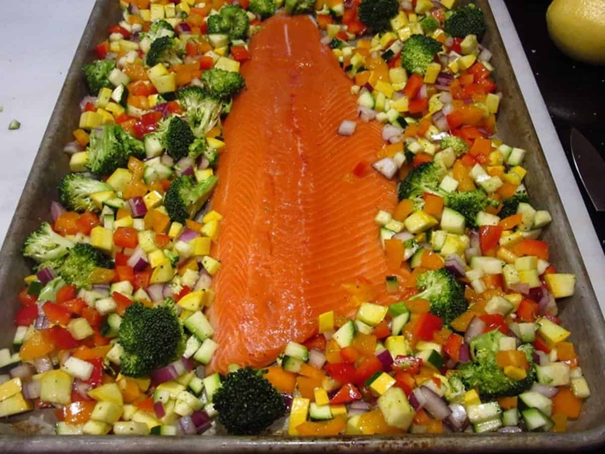 A pink uncooked trout fillet on a sheet pan surrounded by veggies.