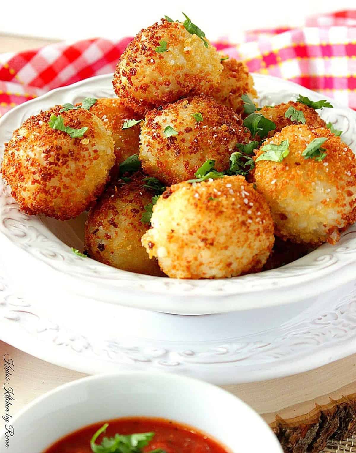 A pretty white bowl filled with cooked arancini balls and a bowl of marinara sauce in the front.