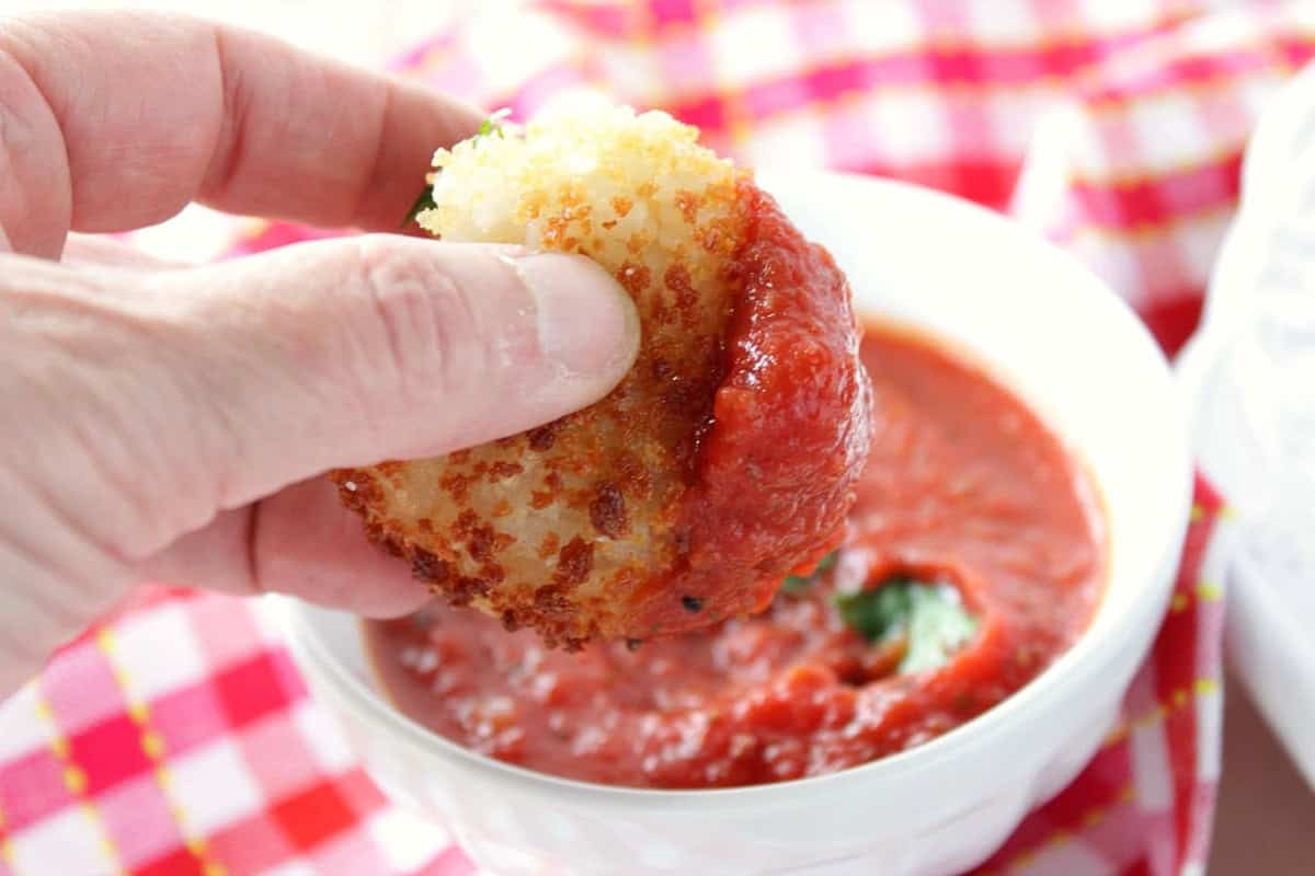 A hand dipping an arancini ball into marinara sauce.