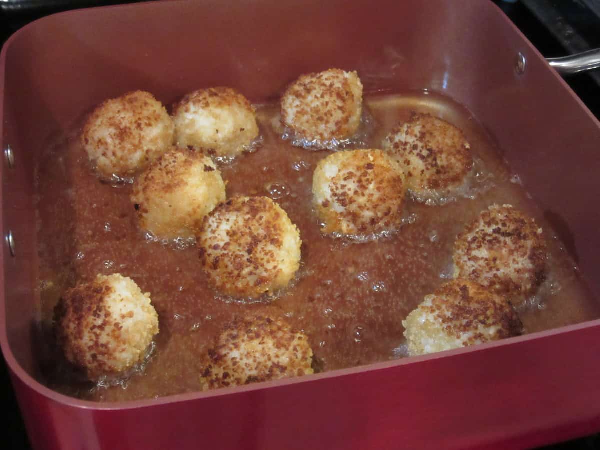 Pan frying arancini balls in oil in a square red pan on the stovetop.